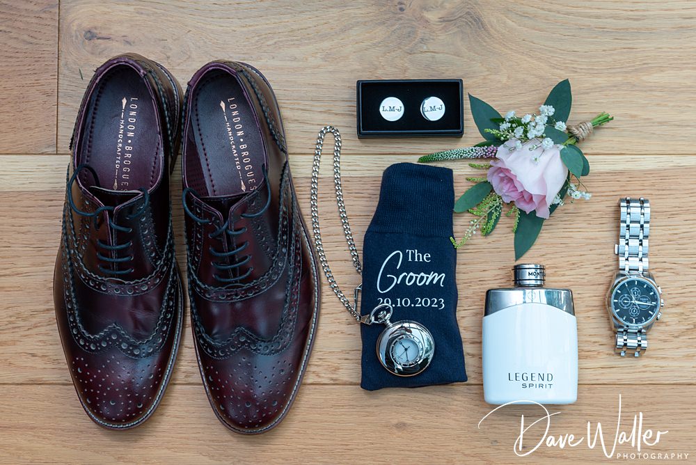 Flat lay of wedding items on wooden floor: polished brown dress shoes, cufflinks, boutonniere, pocket watch, sock with "The Groom 29.10.2023," cologne, and a wristwatch. Captured at Wentbridge House for Georgia & Luke's special day. "Dave Walker Photography" is written in the corner.