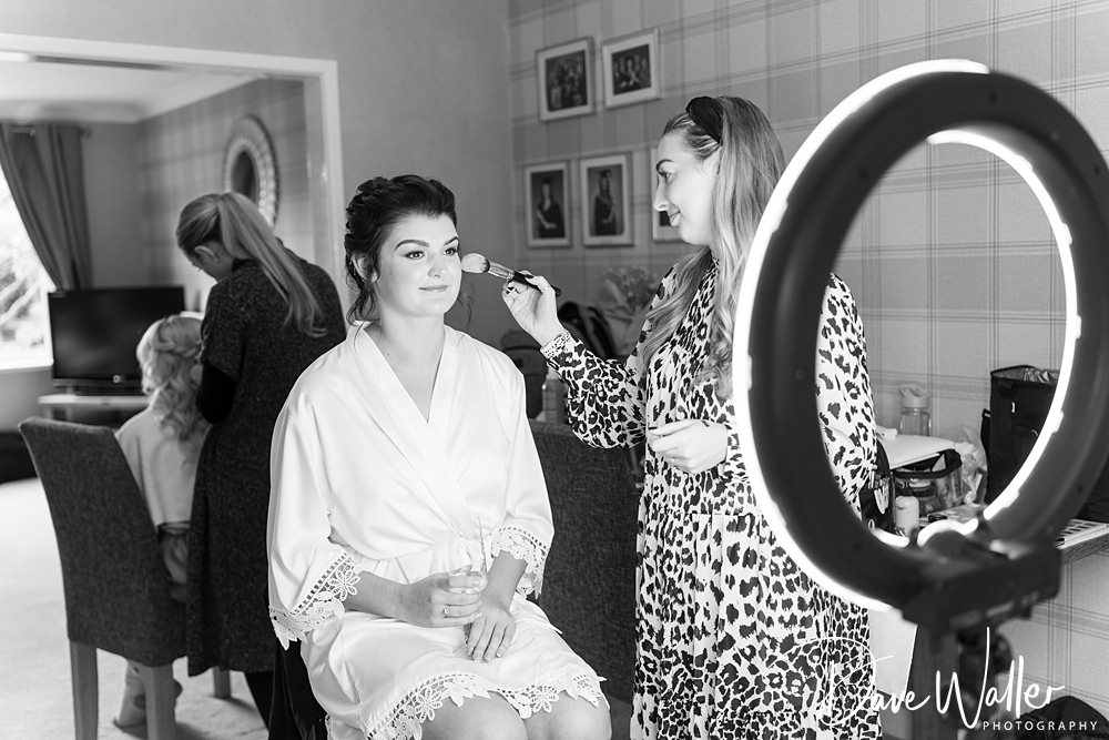 A woman in a robe sits holding a glass, getting her makeup done by a stylist in a patterned outfit. A ring light illuminates them as they prepare for Georgia & Luke's wedding at Wentbridge House, with a TV, several pictures on the wall, and another person in the background.