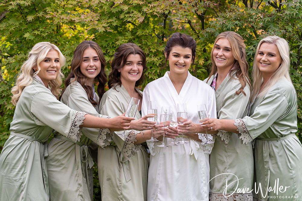 A group of six women in robes stand outdoors at Wentbridge House, smiling and holding champagne glasses. The women in light green robes flank a woman in a white robe, radiating joy amidst the lush greenery surrounding this memorable Wentbridge House Wedding.