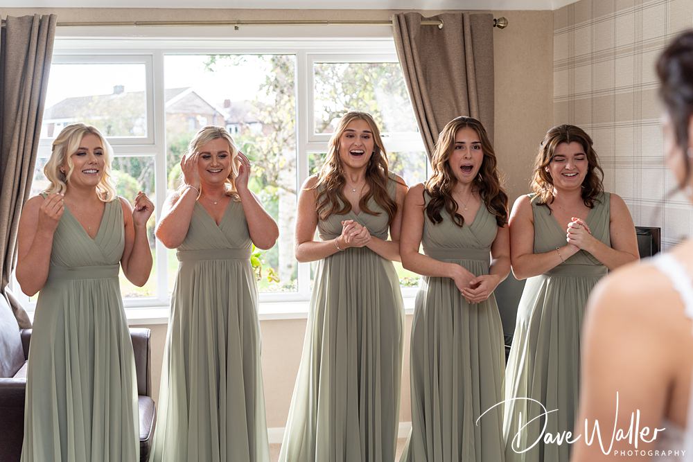 Five bridesmaids in light green dresses react joyfully and excitedly indoors, standing in front of a window with curtains at the Wentbridge House Wedding. The photographer's watermark is visible in the lower right corner.