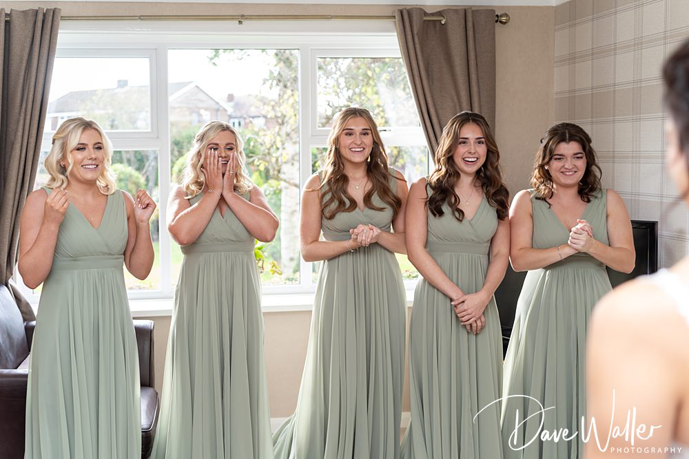 Five bridesmaids in matching sage green dresses express joy as they look toward a bride, who is outside the frame. They're standing in a well-lit room with curtains and a large window in the background, capturing the enchanting atmosphere of Georgia & Luke's wedding at Wentbridge House.