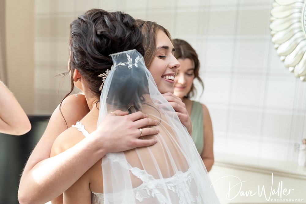 At the Wentbridge House Wedding, a bride adorned with a veil and hair accessory embraces a smiling woman in a heartfelt hug. Another woman beams in the background. The indoor setting, with its plaid patterned walls, captures the joyful essence of Georgia and Luke's special day.