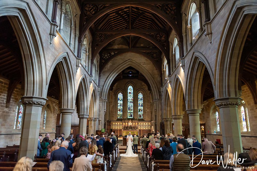 A bride and groom stand at the altar in a large, ornate church with arched ceilings and stained glass windows. Guests fill the pews, seated on either side, witnessing Georgia & Luke's wedding, a beautiful prelude to their Wentbridge House celebration.