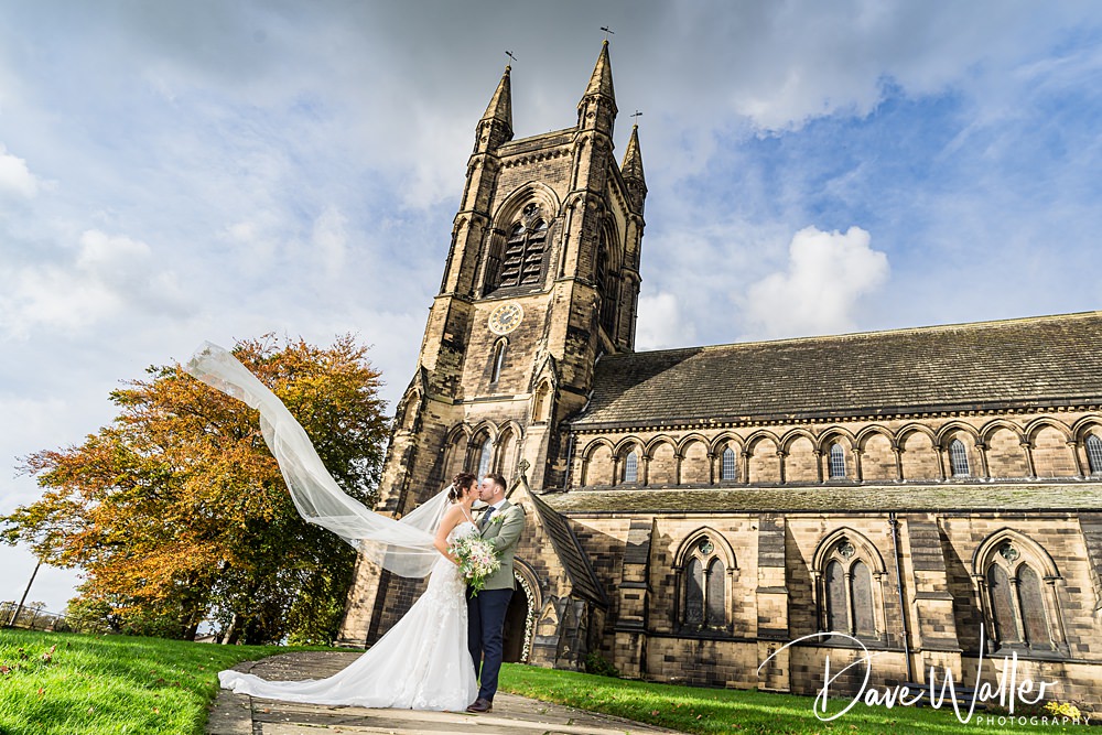Georgia and Luke, in stunning wedding attire, embrace outside the grand stone church of Wentbridge House. The bride's long veil flows elegantly in the wind. The sky is partly cloudy, and an autumn tree with vibrant orange leaves stands nearby, adding a perfect touch to their unforgettable day.