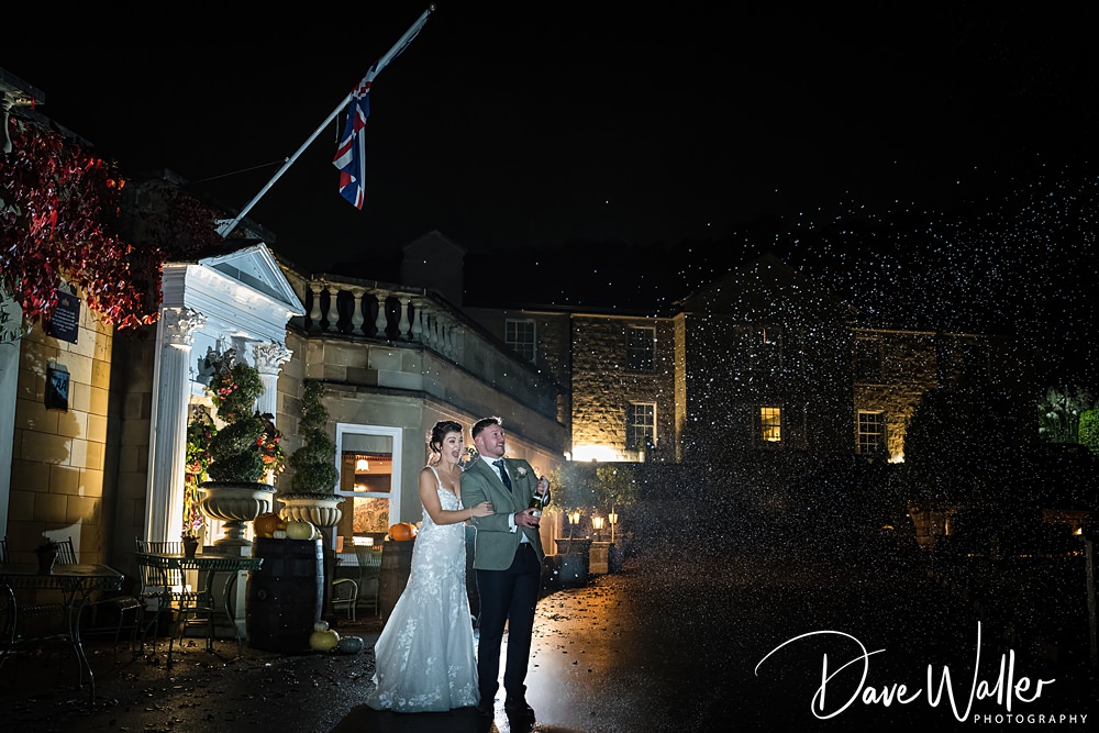 Georgia and Luke, the newlyweds, stand outside Wentbridge House at night, gazing up as confetti falls around them. The scene is beautifully illuminated by building lights, highlighting the elegant architecture and lush greenery. A British flag waves gently above the couple, adding a touch of charm to their wedding celebration.