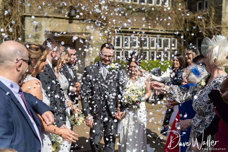 A newlywed couple, Rebecca and Thomas, walk through a crowd of people throwing confetti in celebration outside Holdsworth House. The bride holds a bouquet, and the groom is in a suit under the sunny weather.