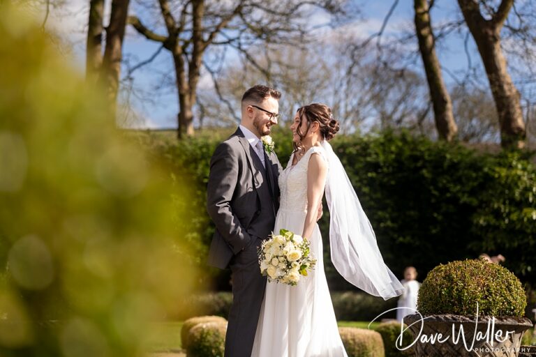 A bride and groom, Rebecca & Thomas, stand close in a garden at Holdsworth House, smiling at each other. The bride holds a bouquet of white flowers, her veil flowing gracefully behind her. Trees and greenery provide a lush backdrop under the sunny sky for this enchanting wedding moment.