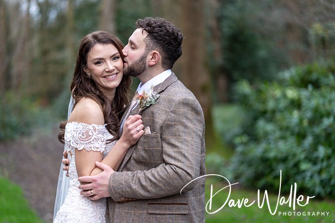 A bride and groom stand outdoors in a dreamy Lincoln setting. The groom, in a brown suit, kisses the smiling bride on the cheek, her off-shoulder lace gown glowing. The blurred forest backdrop adds magic to this moment captured by photographer Dave Waller Photography.
