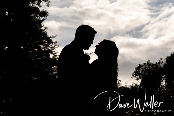 Silhouette of a couple embracing against a cloudy sky backdrop, captured by Lincoln wedding photographer Dave Waller. The scene is beautifully framed by trees, creating an enchanting romantic atmosphere.