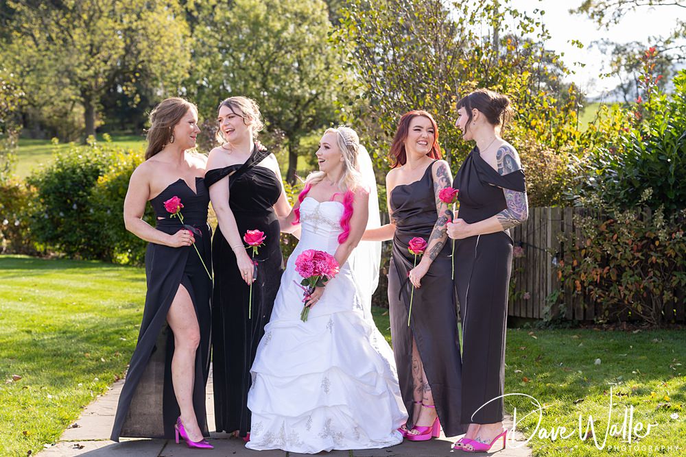 Bride and bridesmaids smiling with pink flowers, outdoors.