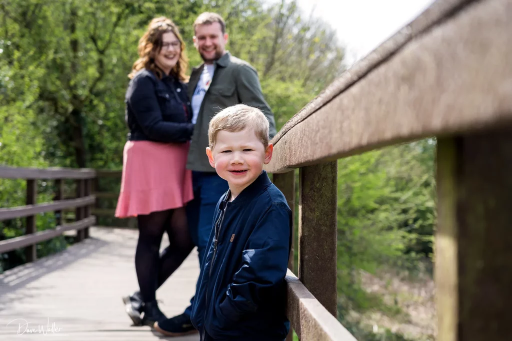 Smiling child on wooden bridge with parents nearby.