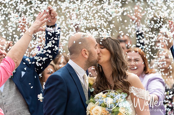 A couple kisses amidst cheering guests throwing flower petals, perfectly captured by Whitley Hall Hotel Wedding Photography. The groom dons a dark suit, while the bride shines in a white dress and holds a bouquet of yellow and white flowers, surrounded by joyful faces and celebration.