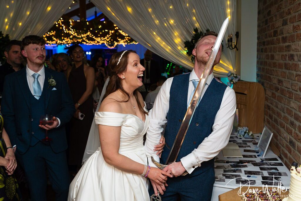 Bride and groom cutting cake with sword, laughing guests.
