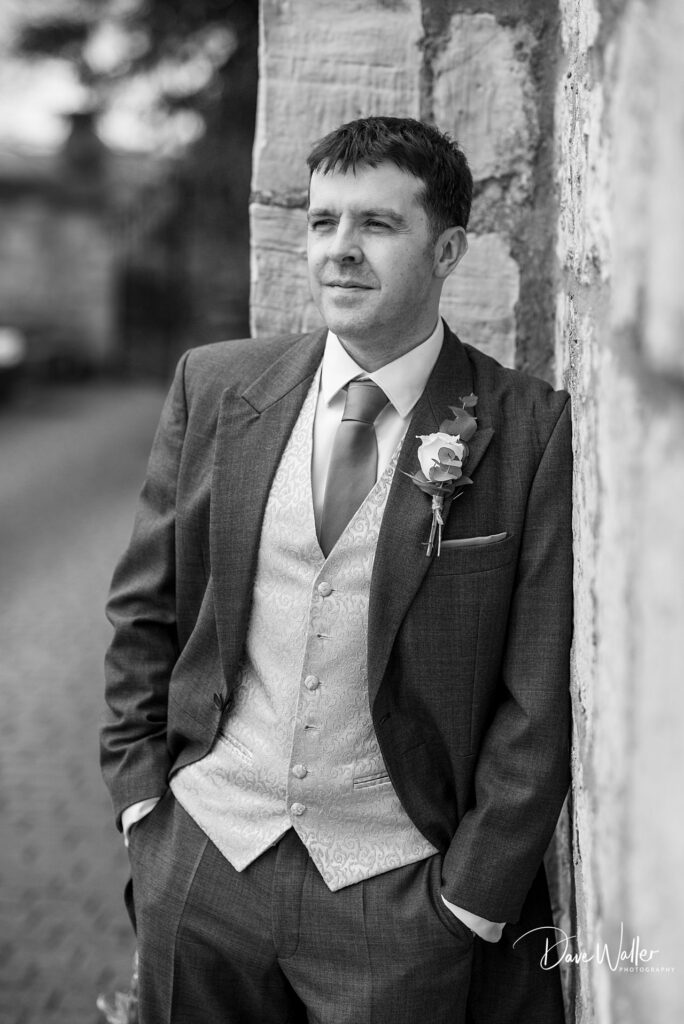 Groom in formal attire leans against stone wall.