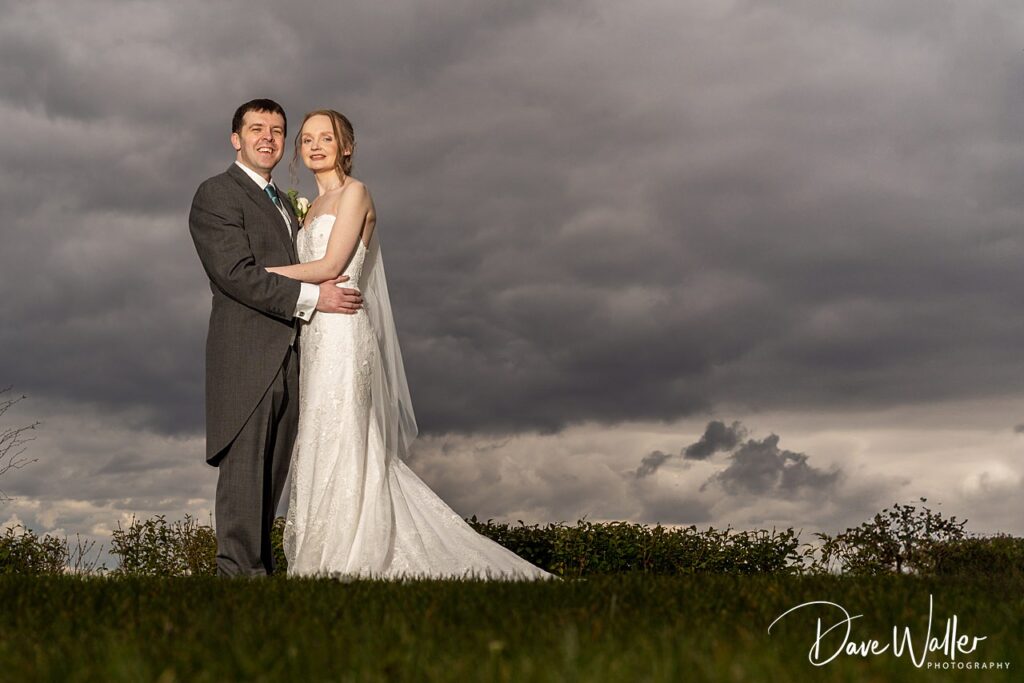 Bride and groom standing together under cloudy sky.