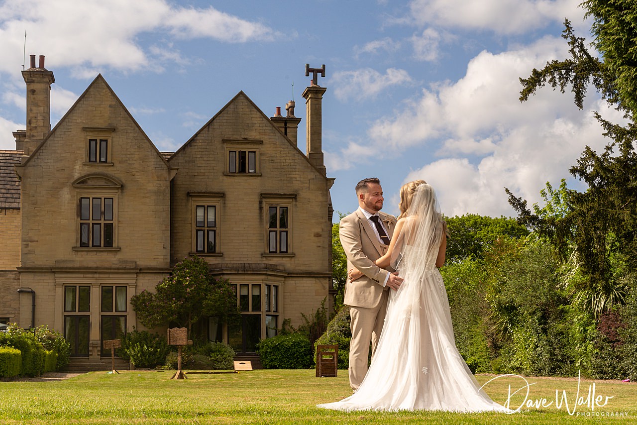 Libby & Jordan stand facing each other on a lawn in front of the grand Bagden Hall Hotel, with clear blue skies and lush greenery surrounding them. The bride wears a white dress and veil; the groom is in a beige suit.