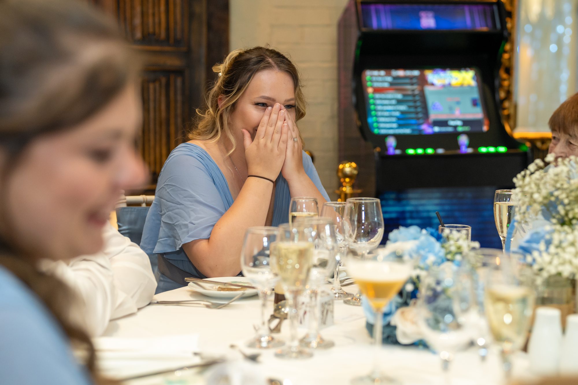 A bridesmaid in blue laughing during the speeches, with the arcade machine behind