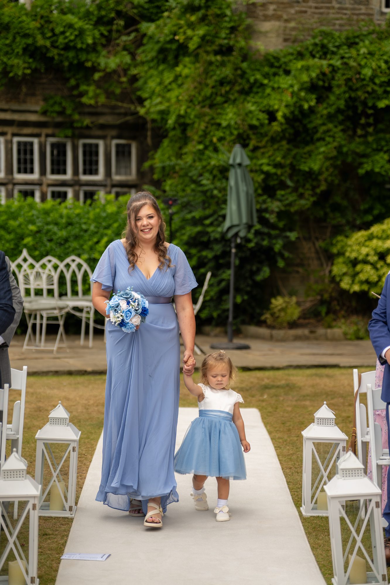A bridesmaid in dusky blue walking down the garden aisle with the flower girl