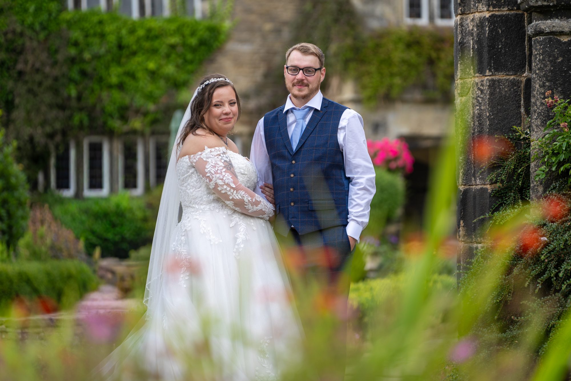 The couple smiling among summer flowers with the manor house windows behind them
