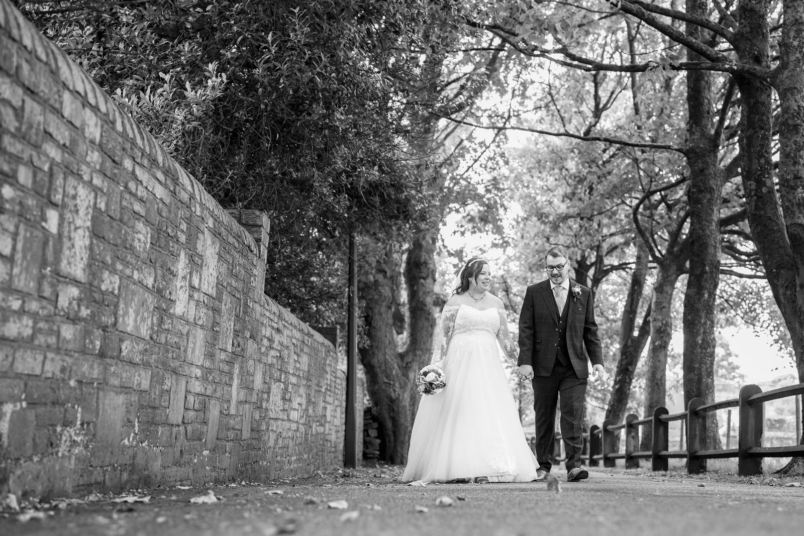 Shannon and Scott walking hand in hand down a tree-lined lane in black and white