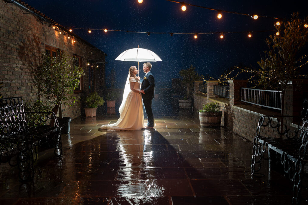 Bride and groom under an umbrella in the rain at an evening wedding